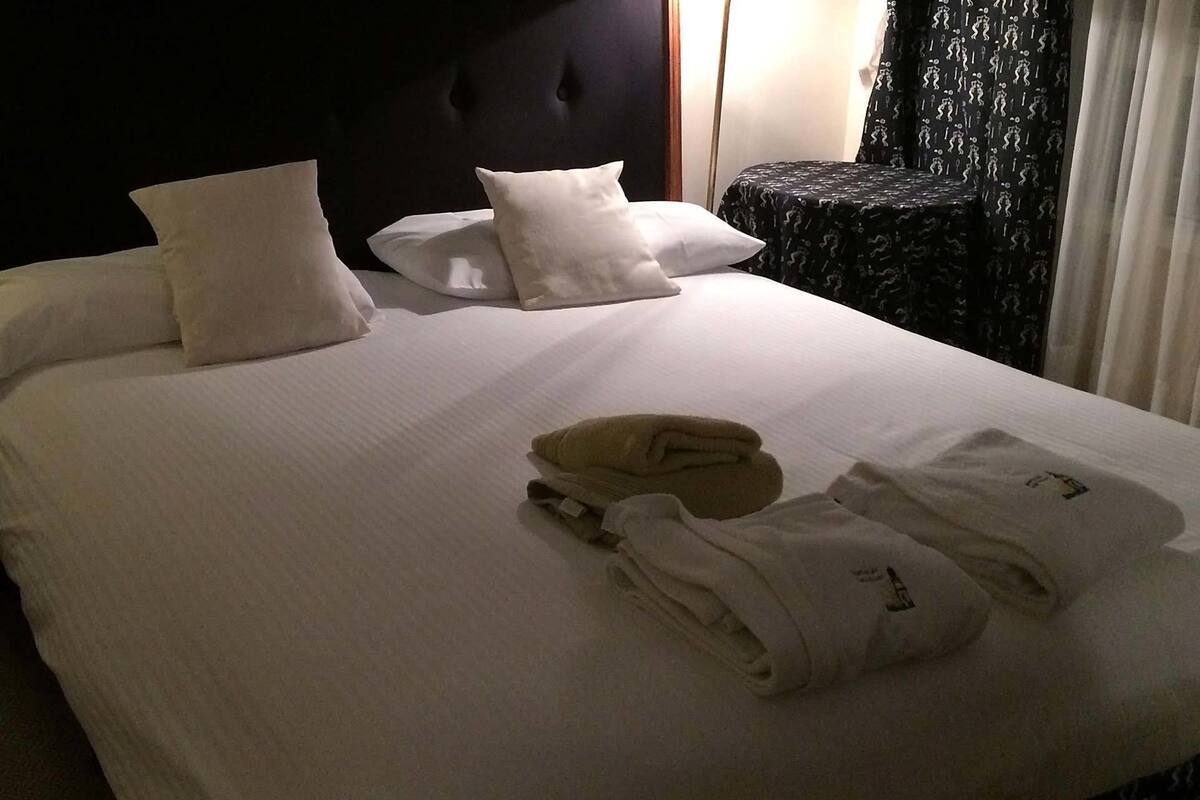 A neatly made bed with white linens and pillows, a folded towel, and a black chair with a patterned cover.