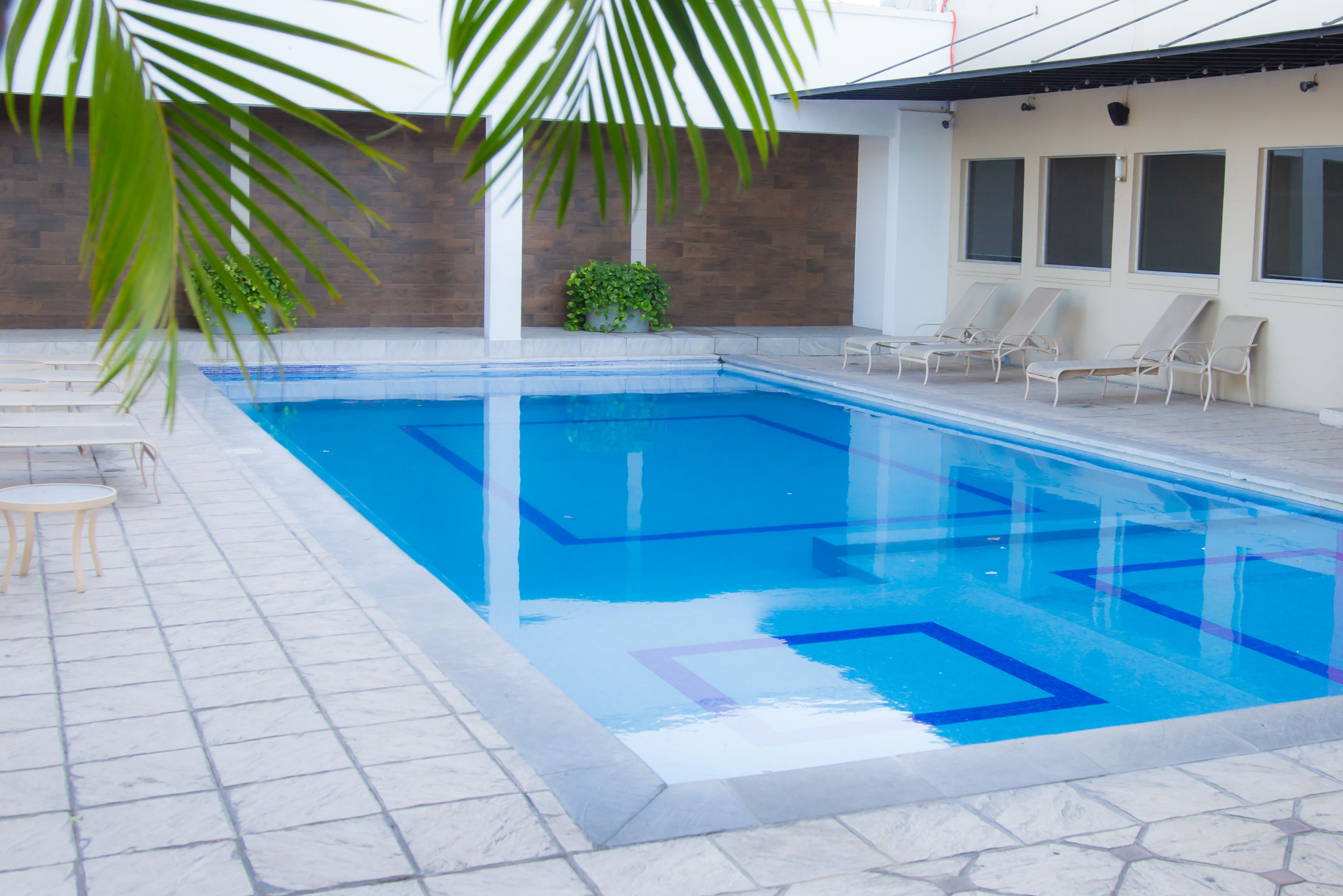 Una piscina rectangular con agua azul cristalina, rodeada por una solana con baldosas. Hay sillas de descanso y una pequeña mesa en la solana.