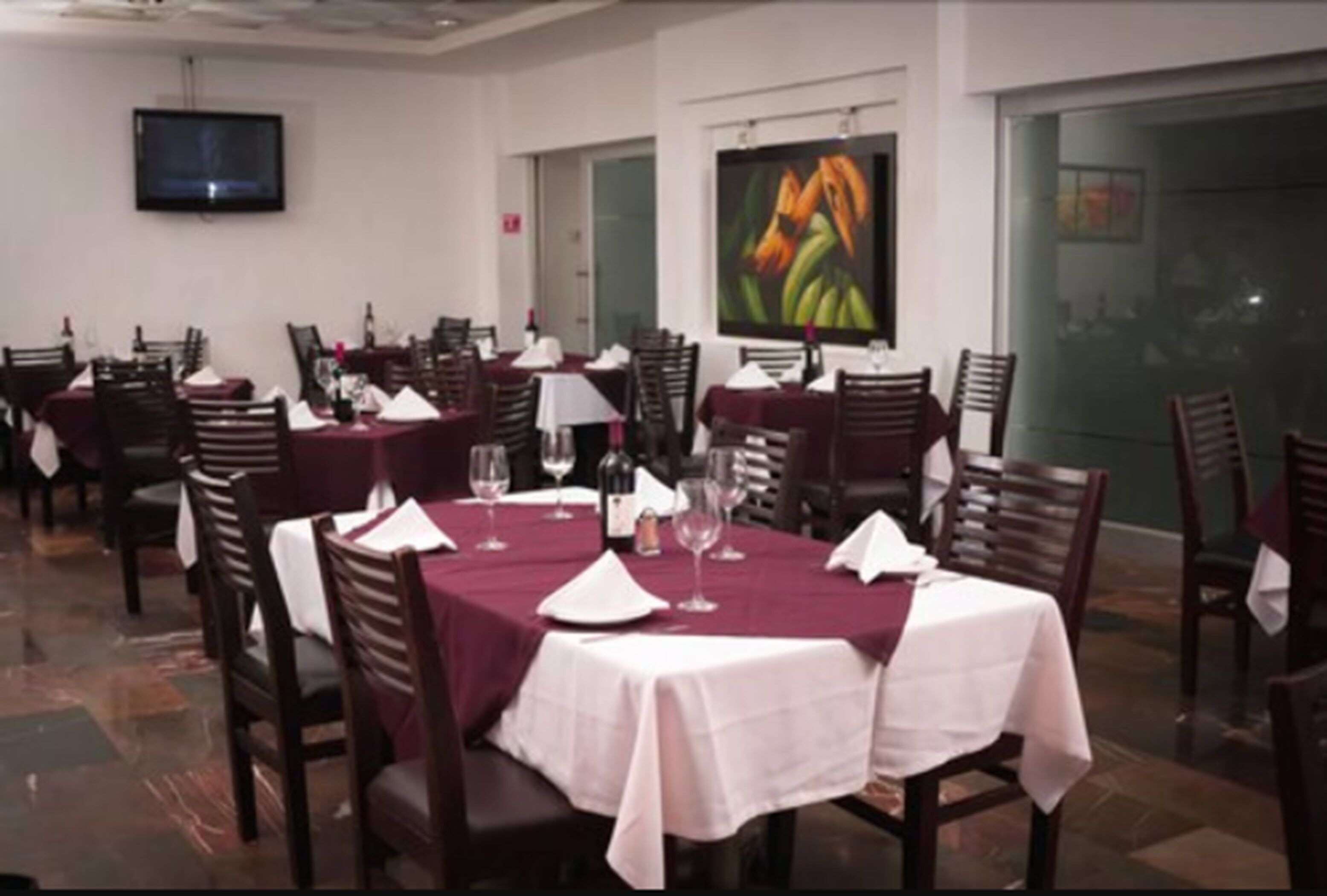 Un espace repas avec des tables préparées pour deux personnes, chacune étant recouverte d’une nappe blanche et agrémentée de verres à vin. Un téléviseur est fixé au mur et un tableau est accroché au-dessus des tables.
