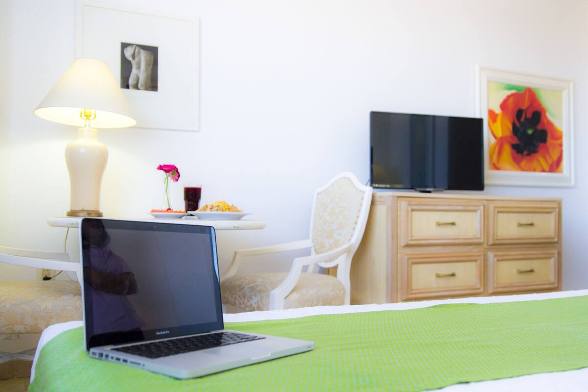 Habitación de hotel con cama, laptop sobre la cama, televisor en un soporte, lámpara, silla, cómoda y una obra de arte en la pared.