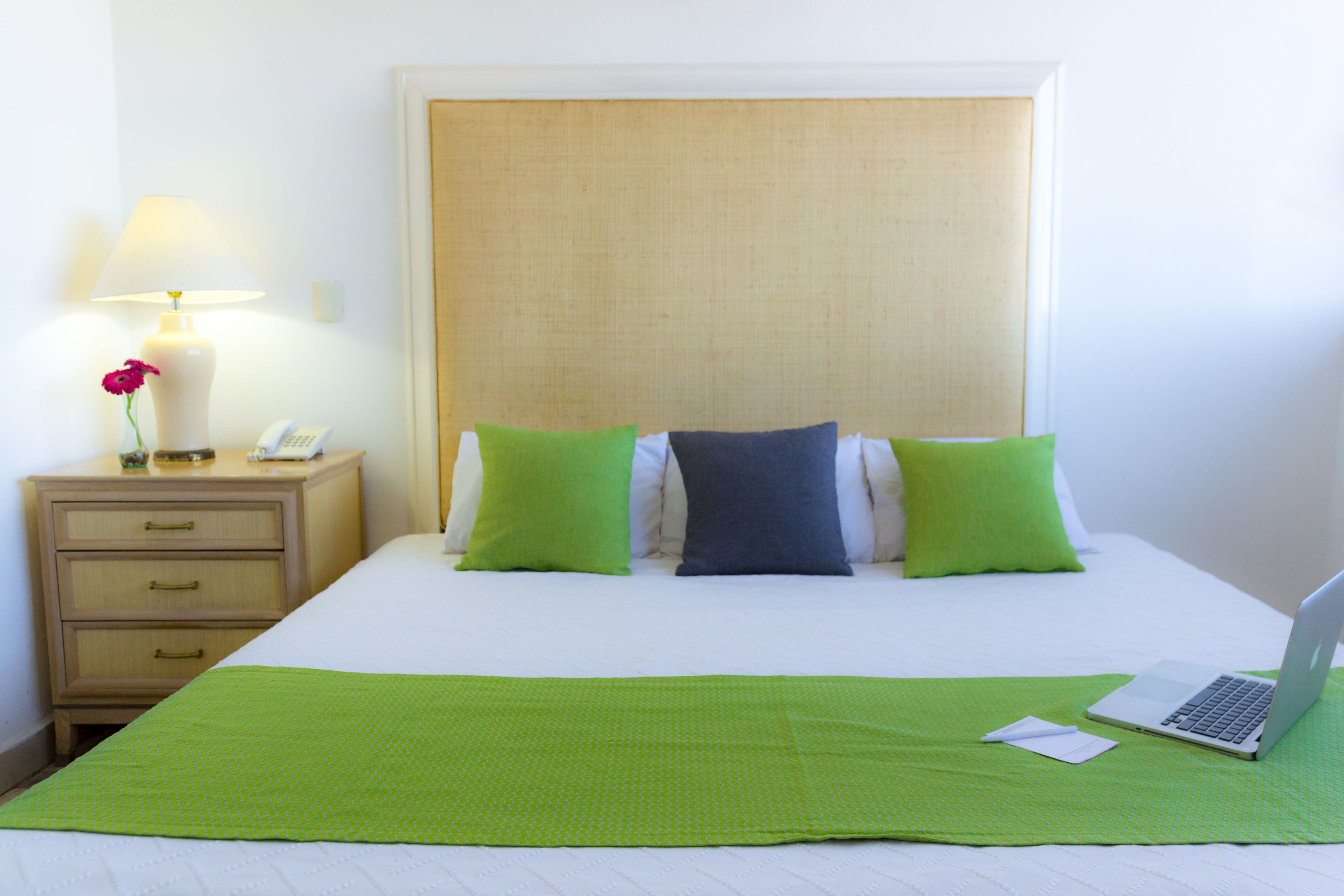 Una cama bien tendida con una colcha verde, una computadora portátil sobre la cama y una mesita de noche con una lámpara y un florero con flores.