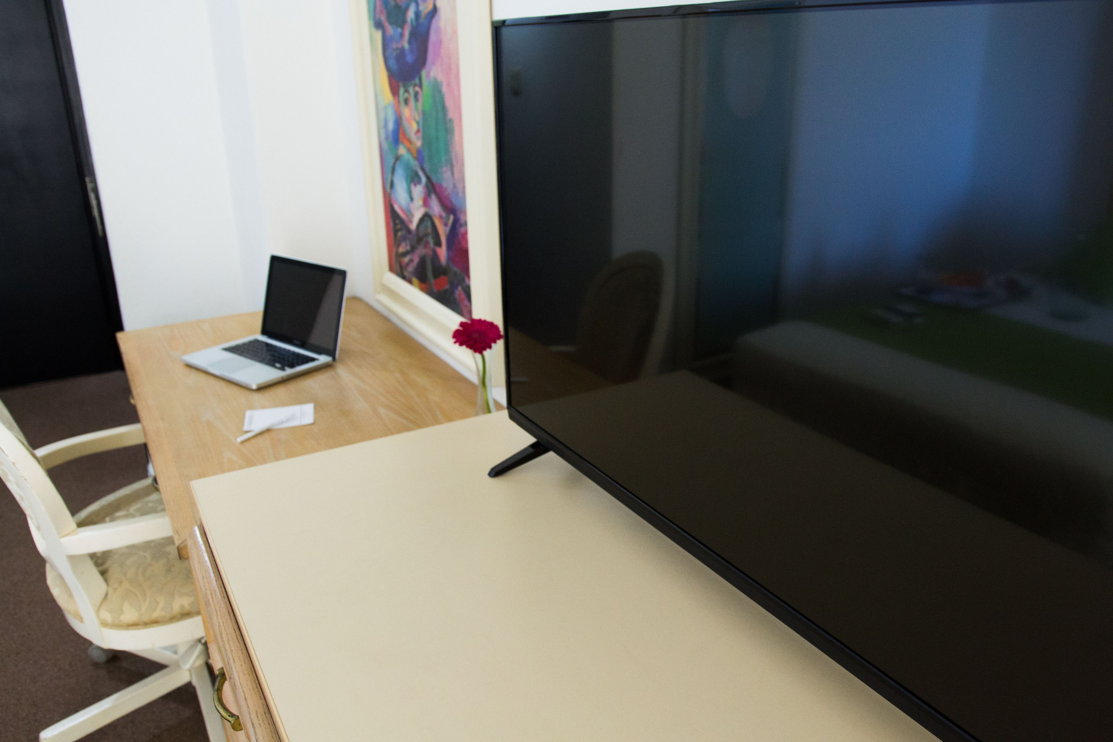 Una habitación con un escritorio de madera, una computadora portátil, un televisor de pantalla plana grande y un cuadro llamativo en la pared.