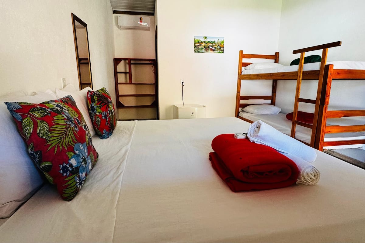 A room with a bunk bed, a single bed with a red blanket, and a small table with a lamp.
