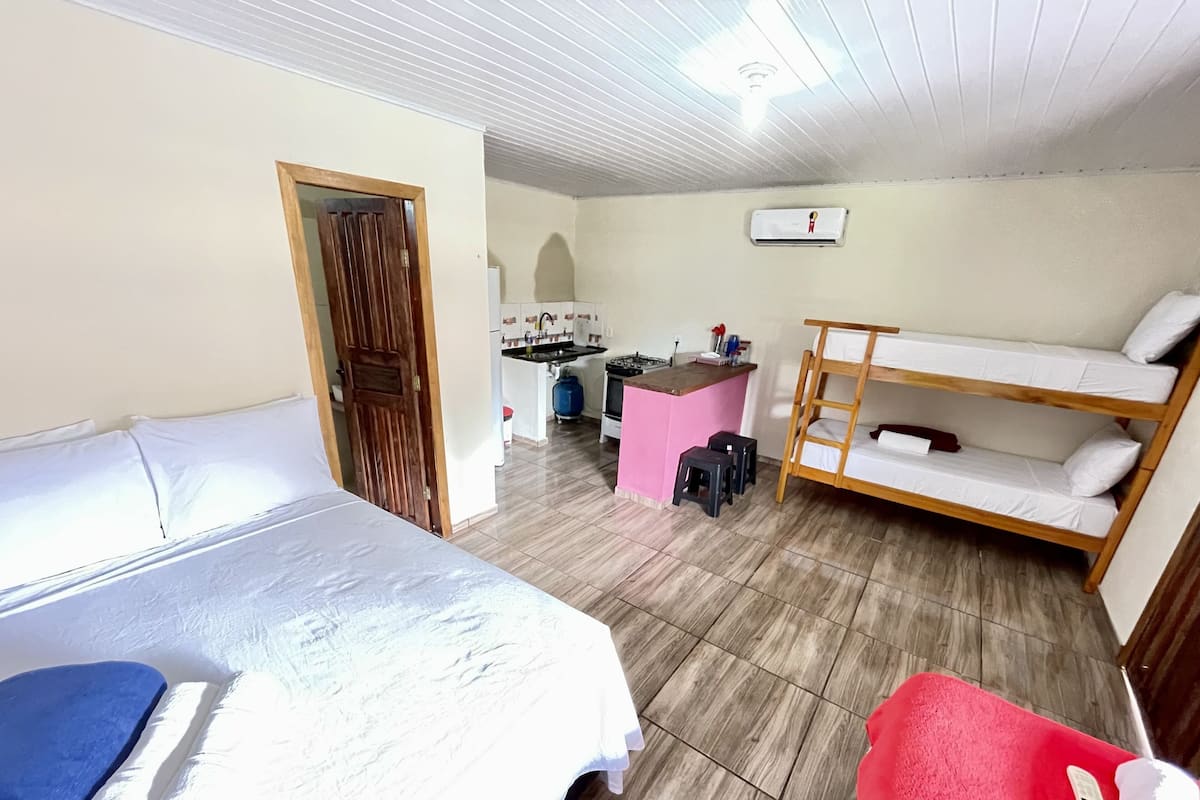 A room with a bunk bed, a single bed, a kitchenette, and a door leading to another room.
