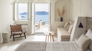 A bedroom with a bed, a chair, a sofa, a small table, and a view of the sea through open doors. - Enigma Suites (Santorini)