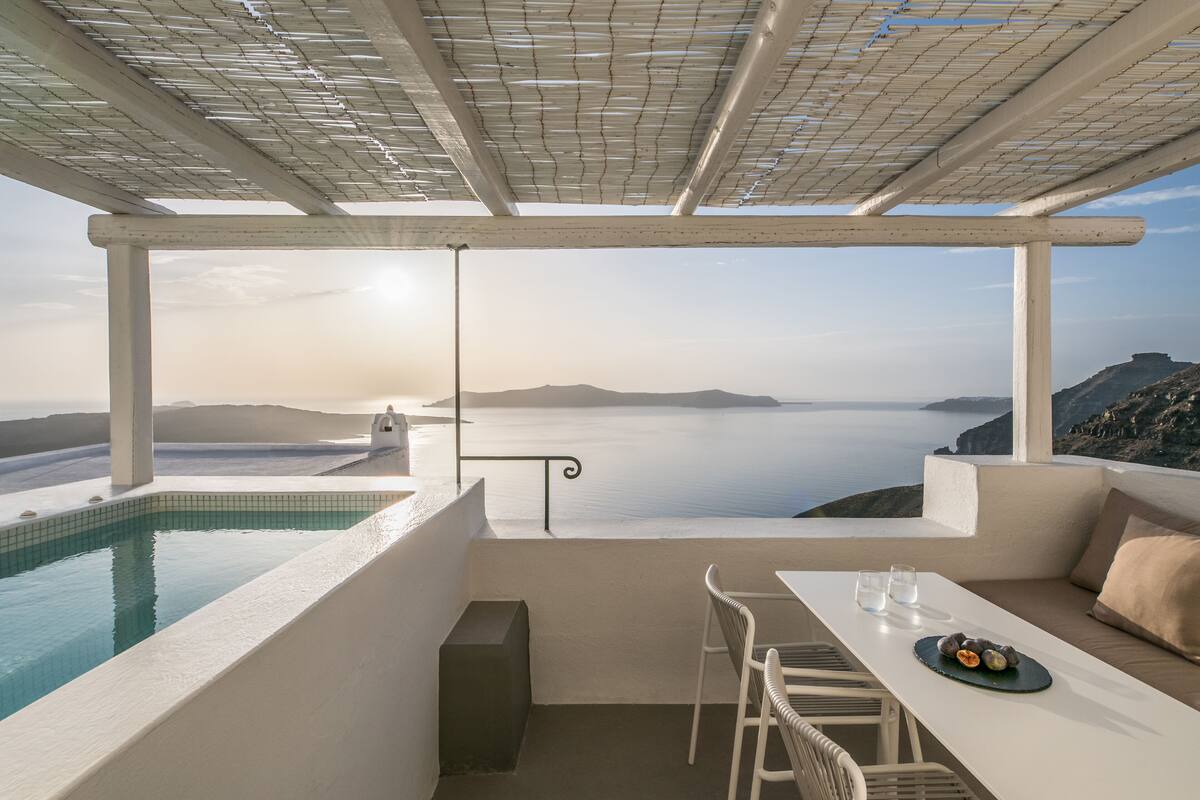 Um terraço com piscina, mesa com copos e prato de comida, e vista para o mar e montanhas.