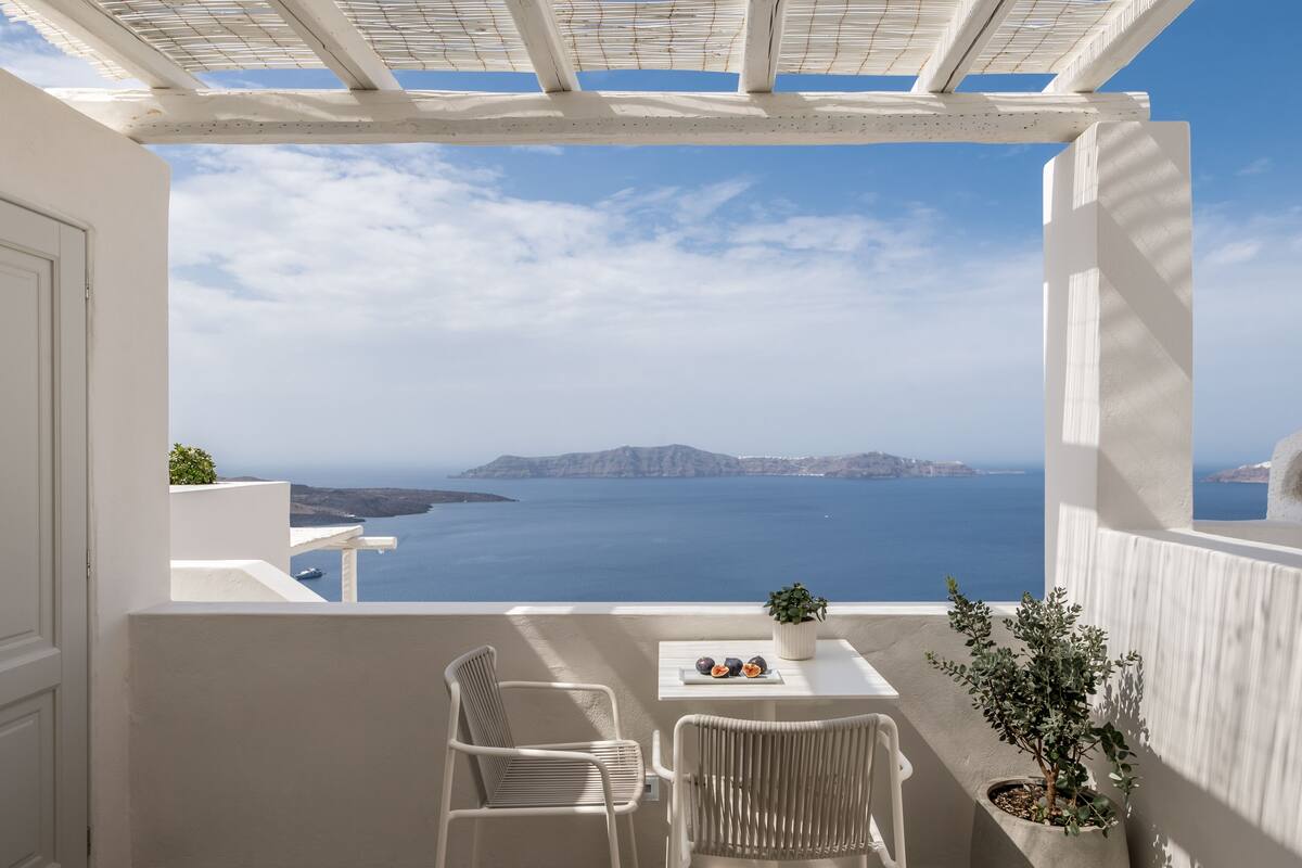 Uma varanda com cadeiras de vime branco, uma pequena mesa com um vaso de planta e vista para o mar e ilhas.
