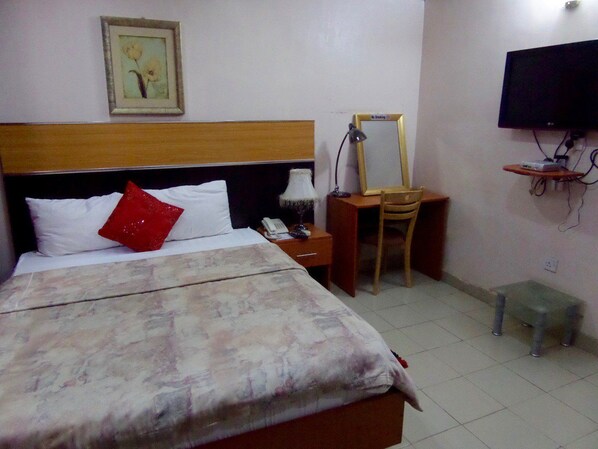 Habitación de hotel con una cama, una mesita de noche con lámpara, una silla, un televisor montado en la pared y un taburete pequeño.