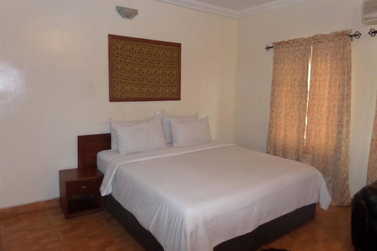 A bedroom with a single bed, a nightstand, a wall-mounted light, and a window with curtains.