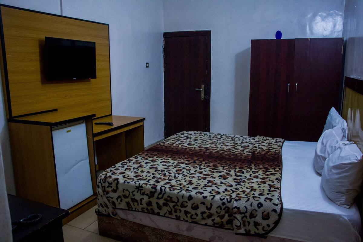 A bedroom with a bed, a television mounted on a wooden panel, a wardrobe, and a small desk.