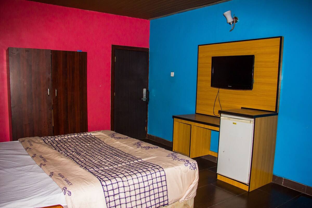A bedroom with a bed, a wooden wardrobe, a small white fridge, a wooden desk, and a television mounted on the wall.