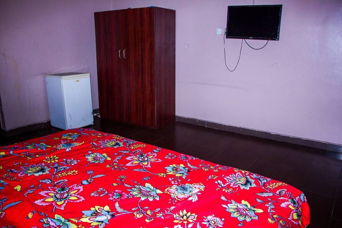 A room with a bed, a wardrobe, a small white refrigerator, and a television mounted on the wall.