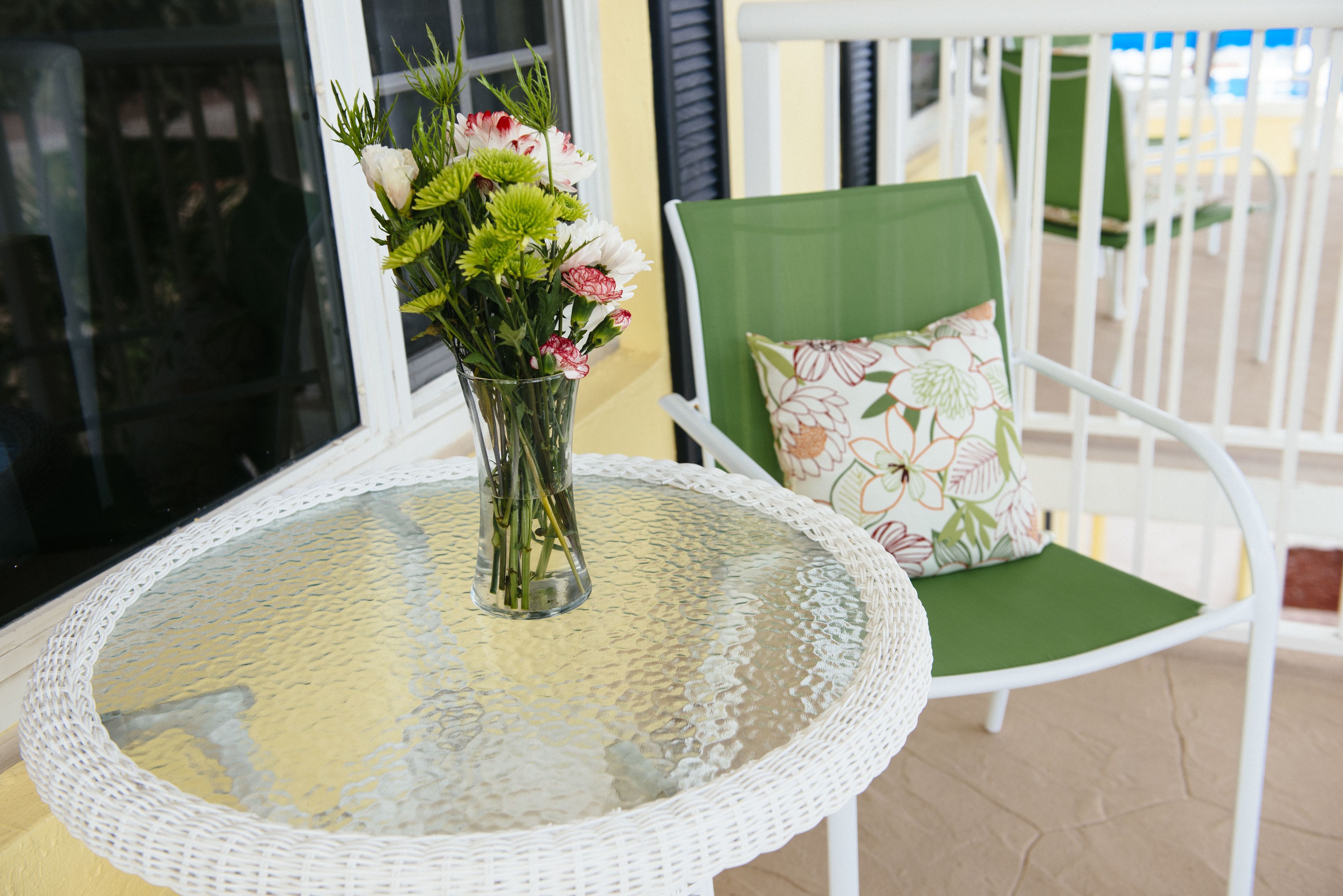 Een ronde glazen tafel met een witte rieten stoel ernaast, een vaas met bloemen en een raam met jaloezieën.