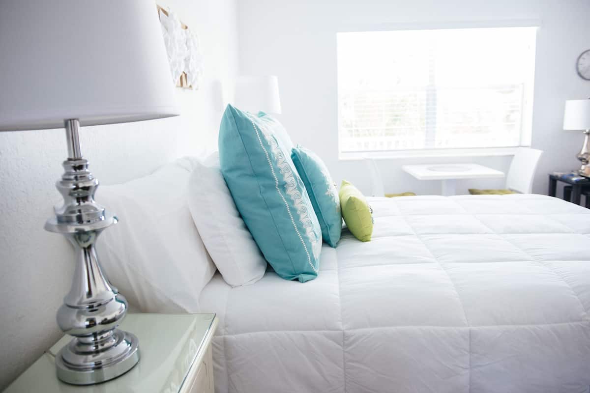 Uma cama bem arrumada com almofadas brancas e azul-turquesa, um abajur e uma janela com vista para a cidade.
