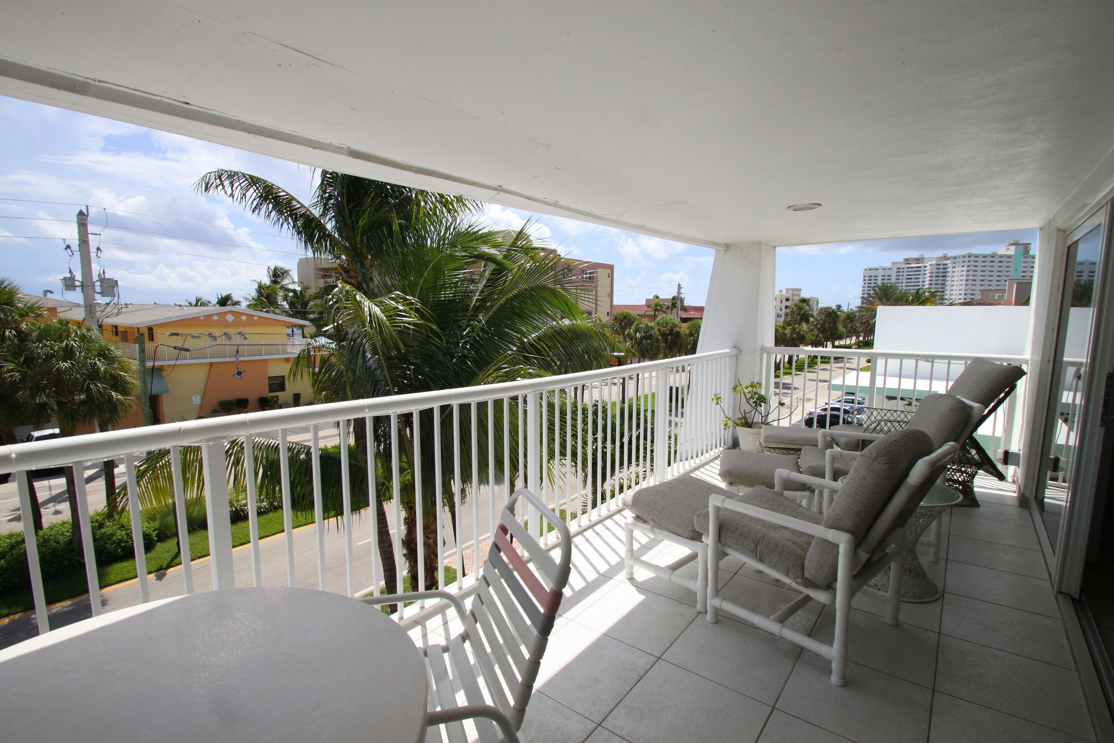 A balcony with white railings, a table, and two chairs. There are palm trees and buildings in the background.