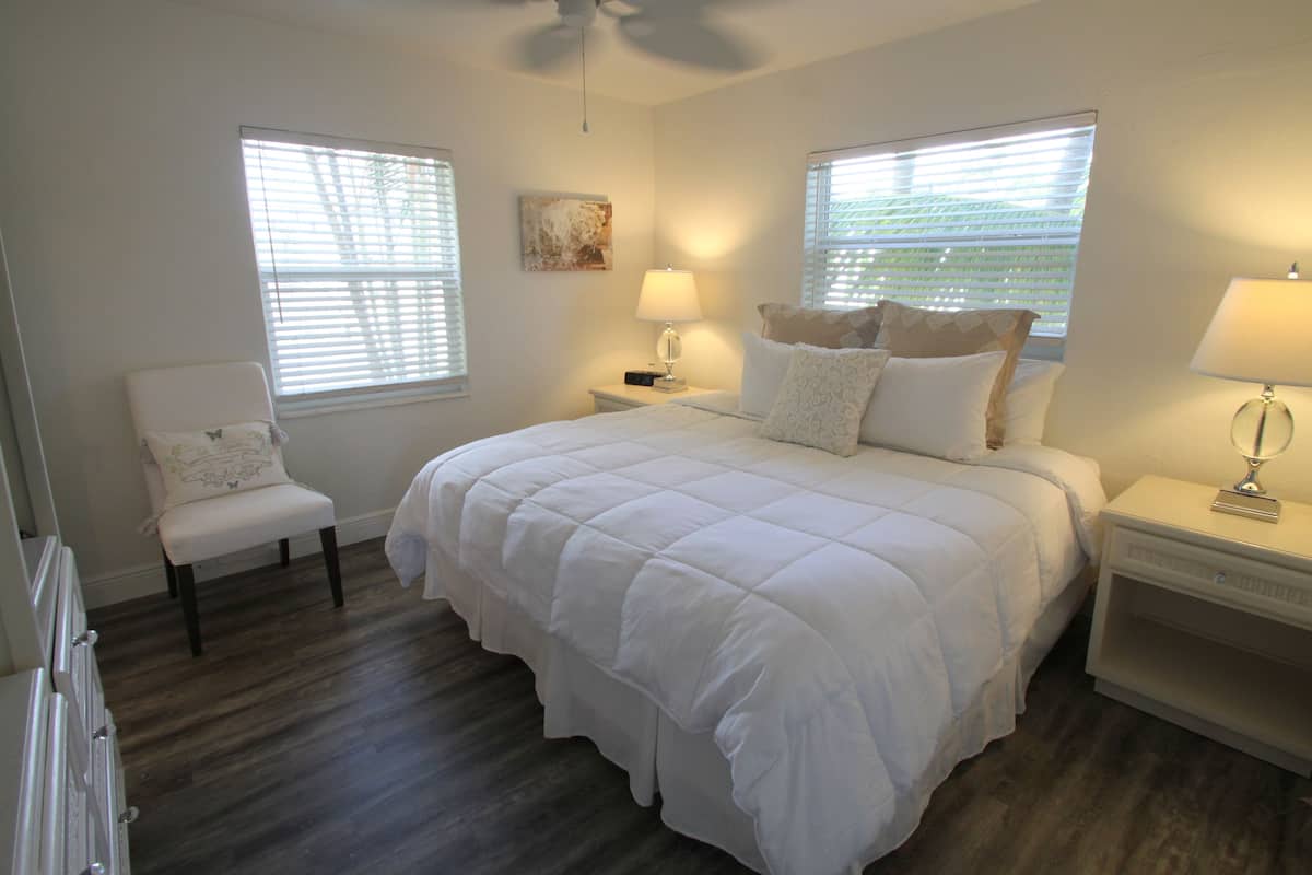 Quarto com cama de casal, duas mesas de cabeceira com luminárias, uma cadeira e duas janelas com persianas.