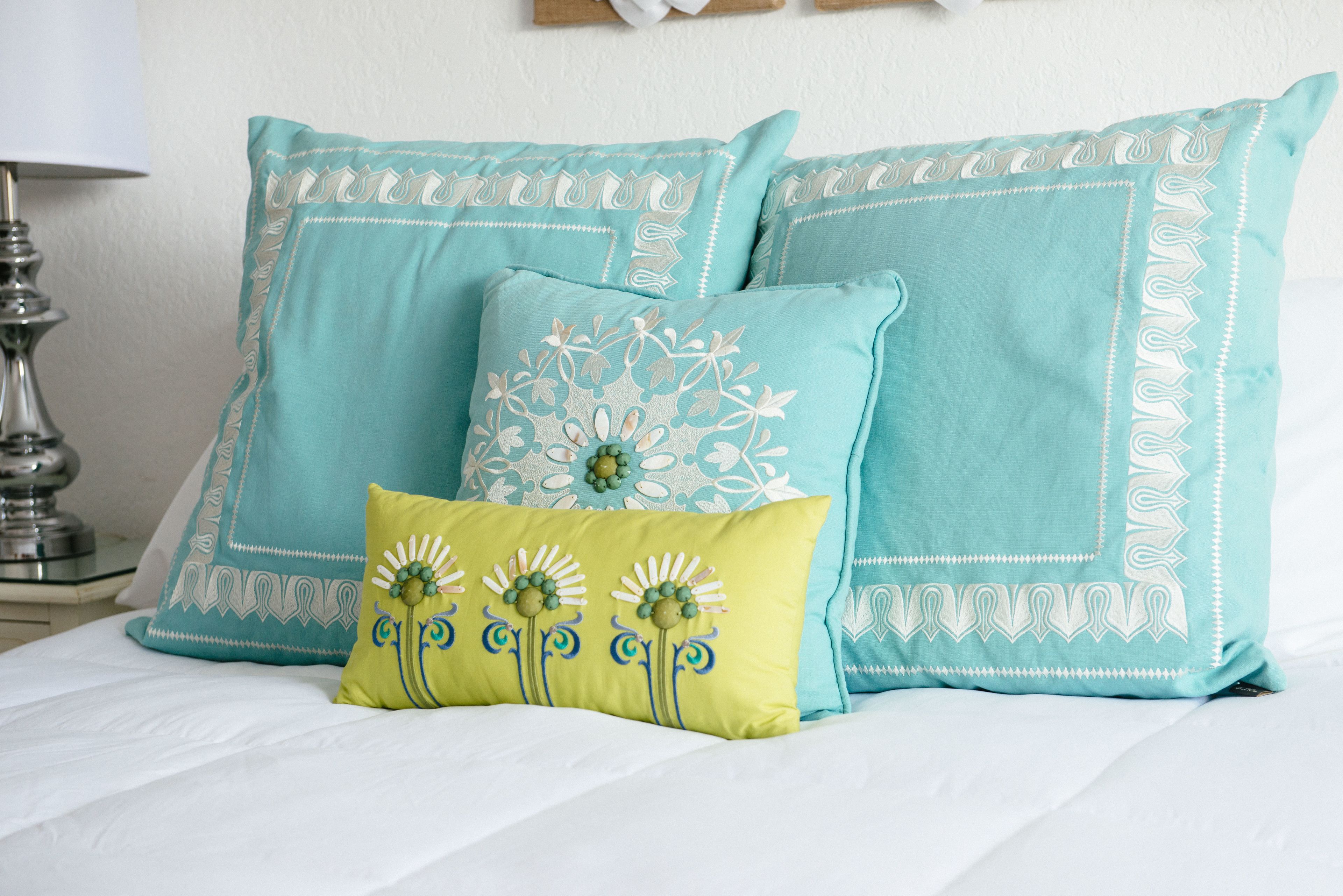 A bed with three pillows, two large teal pillows with white patterns and one smaller yellow pillow with floral embroidery.