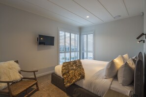 Ein Schlafzimmer mit einem Bett, einem Sessel, einem Fernseher und großen Fenstern mit Jalousien.