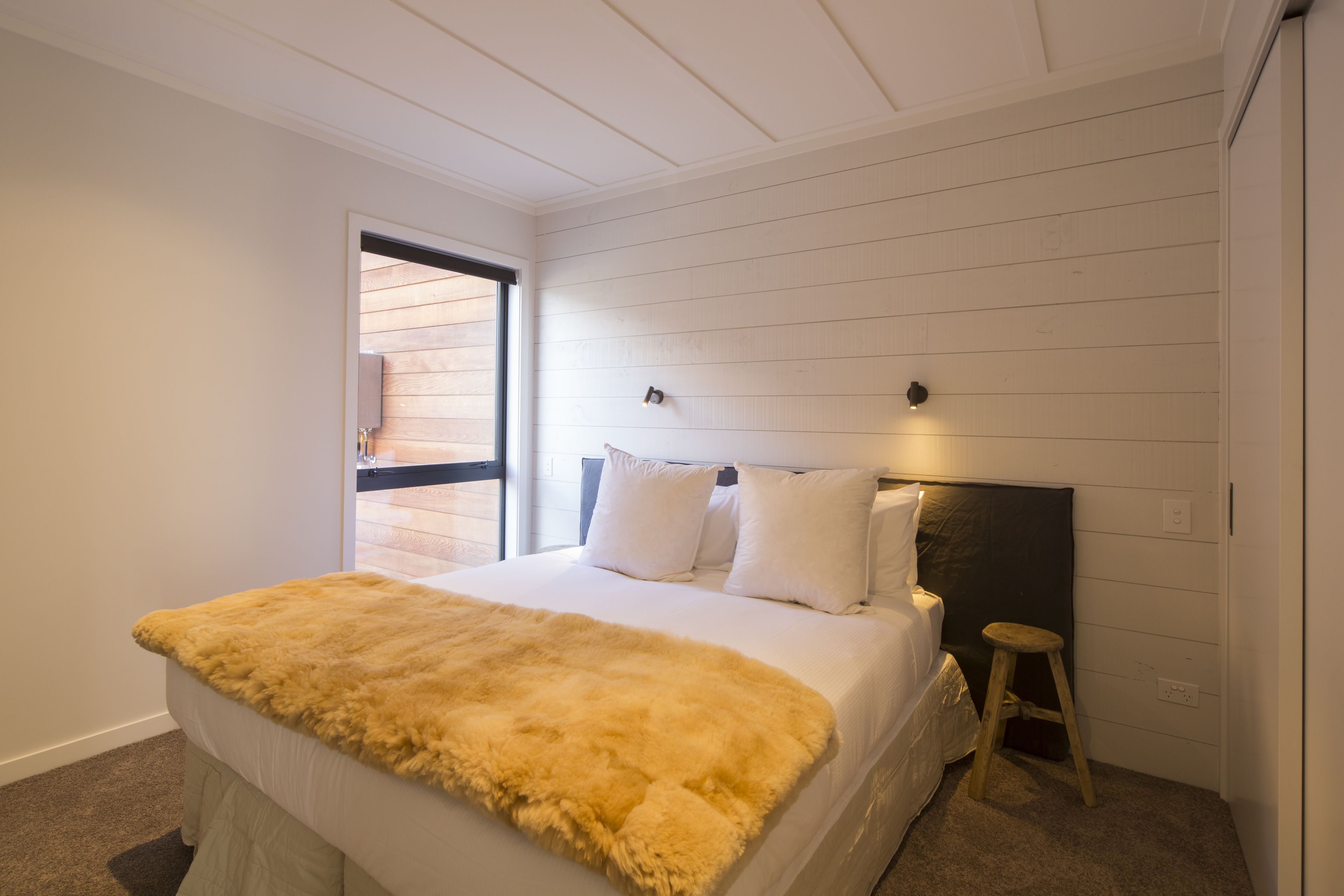 A bedroom with a bed, a wooden headboard, a small stool, and a window with blinds.