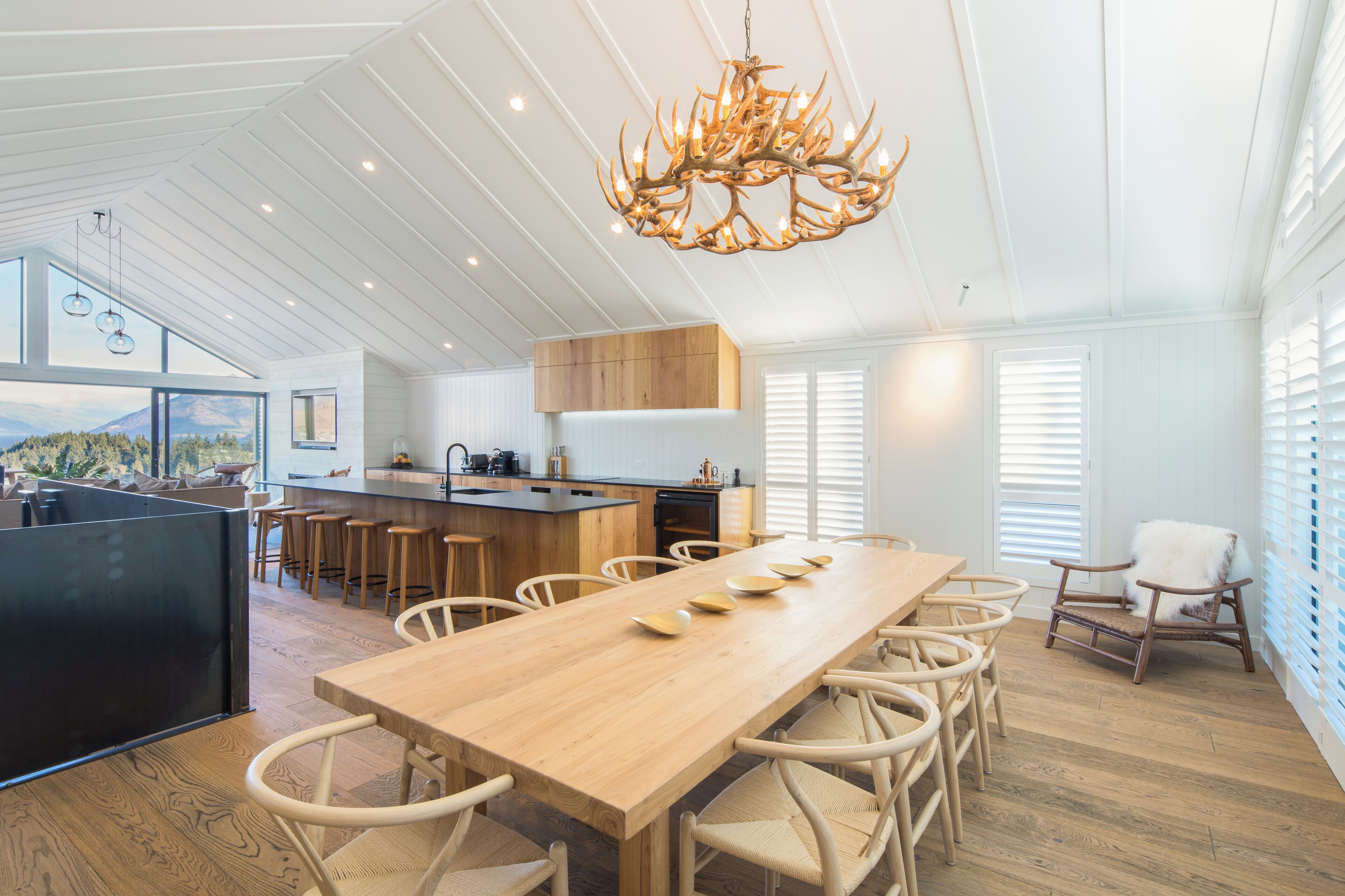 A spacious kitchen with a long wooden table and chairs, a modern chandelier, and a view of the mountains through large windows.