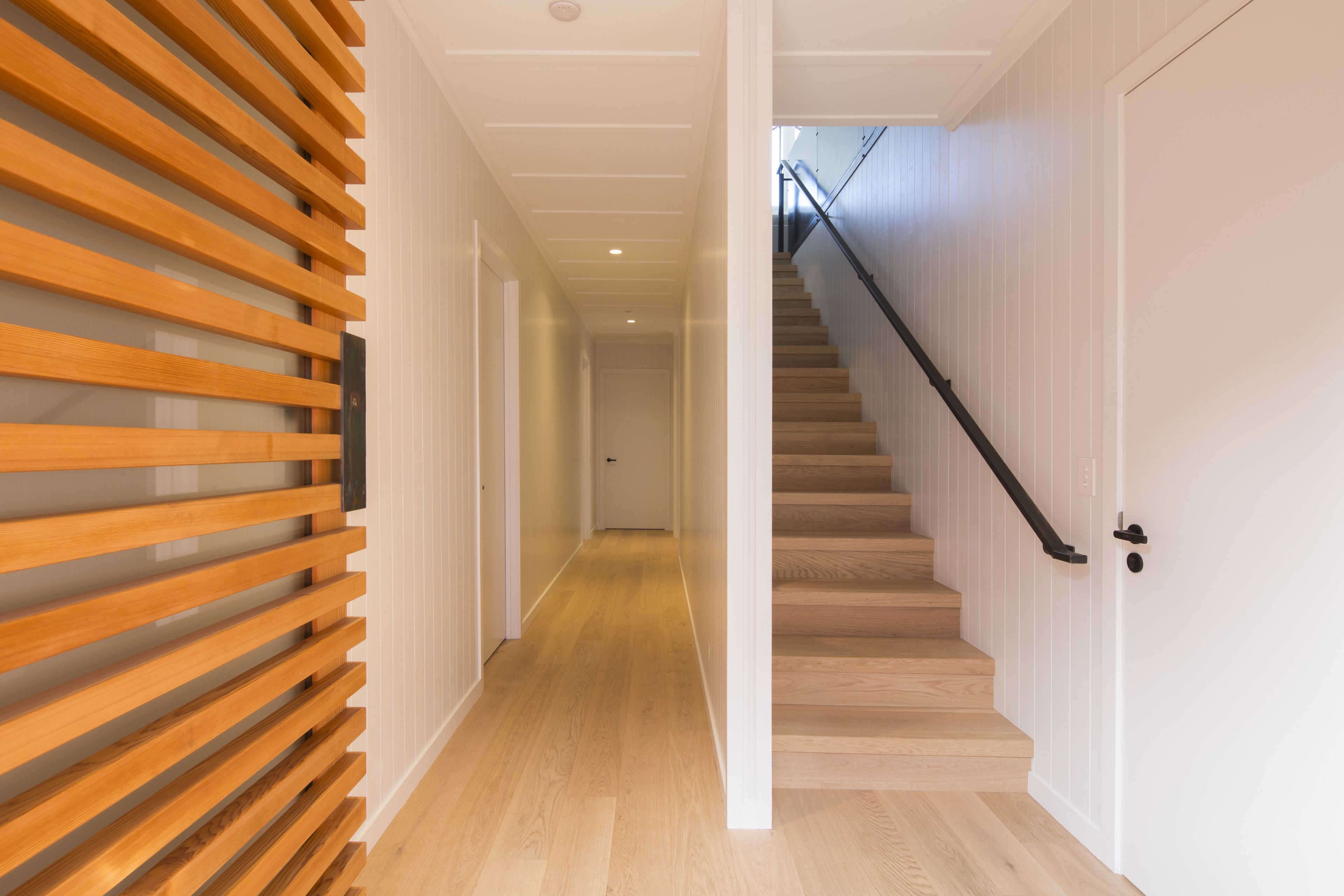 A hallway with wooden slats on one side and a staircase with a black railing leading to a door.