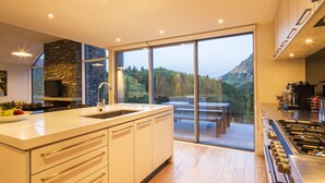 A modern kitchen with a large island, a stone accent wall, and a view of a deck and forested mountains. - Riverview Retreat (Arthur's Point)