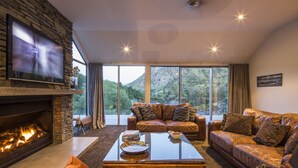 A modern living room with a stone fireplace, leather sofas, a flat-screen TV, and large windows offering a mountain view. - Riverview Retreat (Arthur's Point)