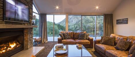 A modern living room with a stone fireplace, leather sofas, a flat-screen TV, and large windows offering a mountain view.