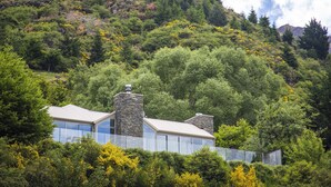 A modern house with a stone chimney, surrounded by dense green forest and a mountain in the background. - Riverview Retreat (Arthur's Point)