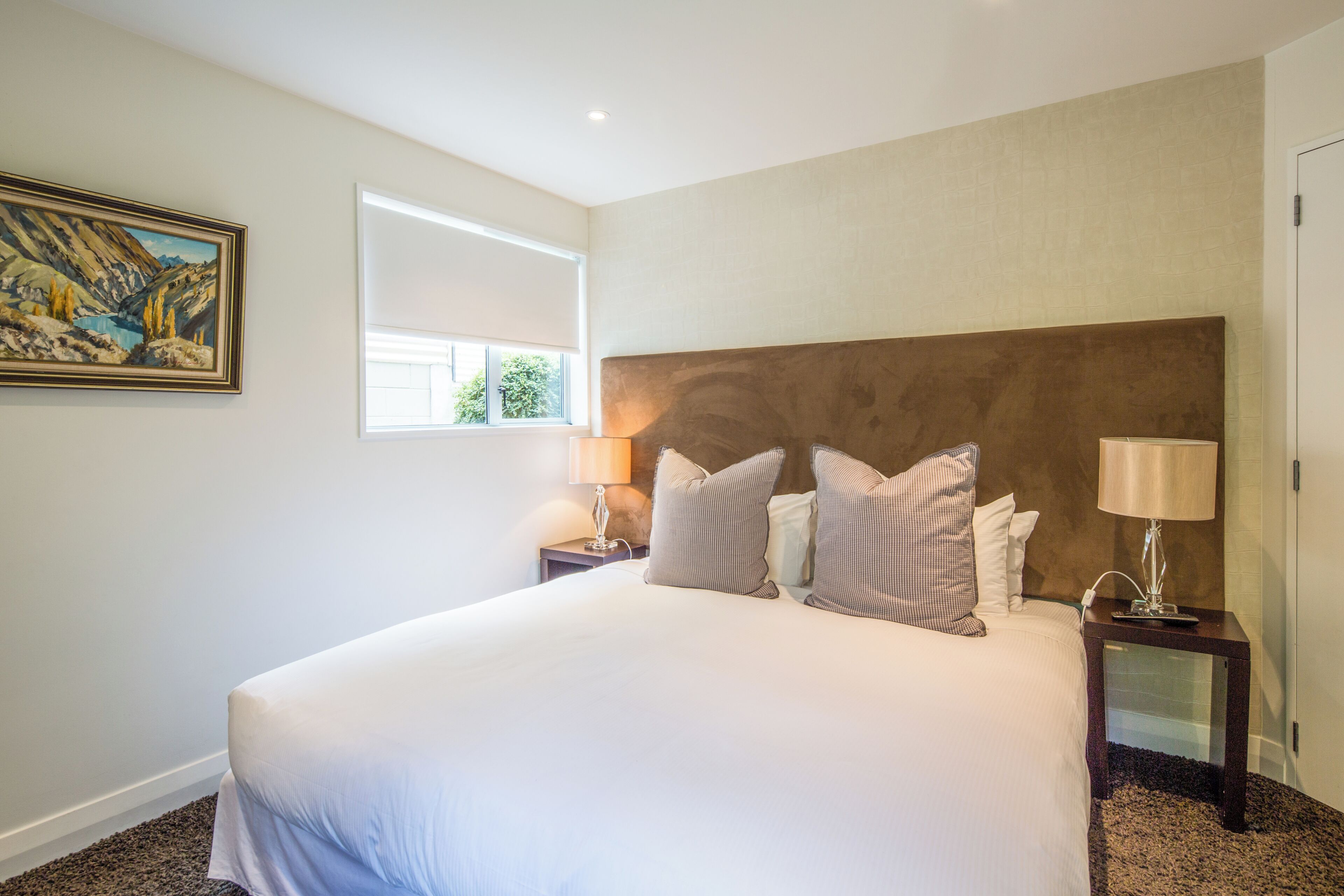 A large bed with a headboard, two bedside tables with lamps, a painting on the wall, and a window providing natural light.