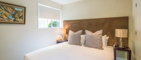 A large bed with a headboard, two bedside tables with lamps, a painting on the wall, and a window providing natural light.
