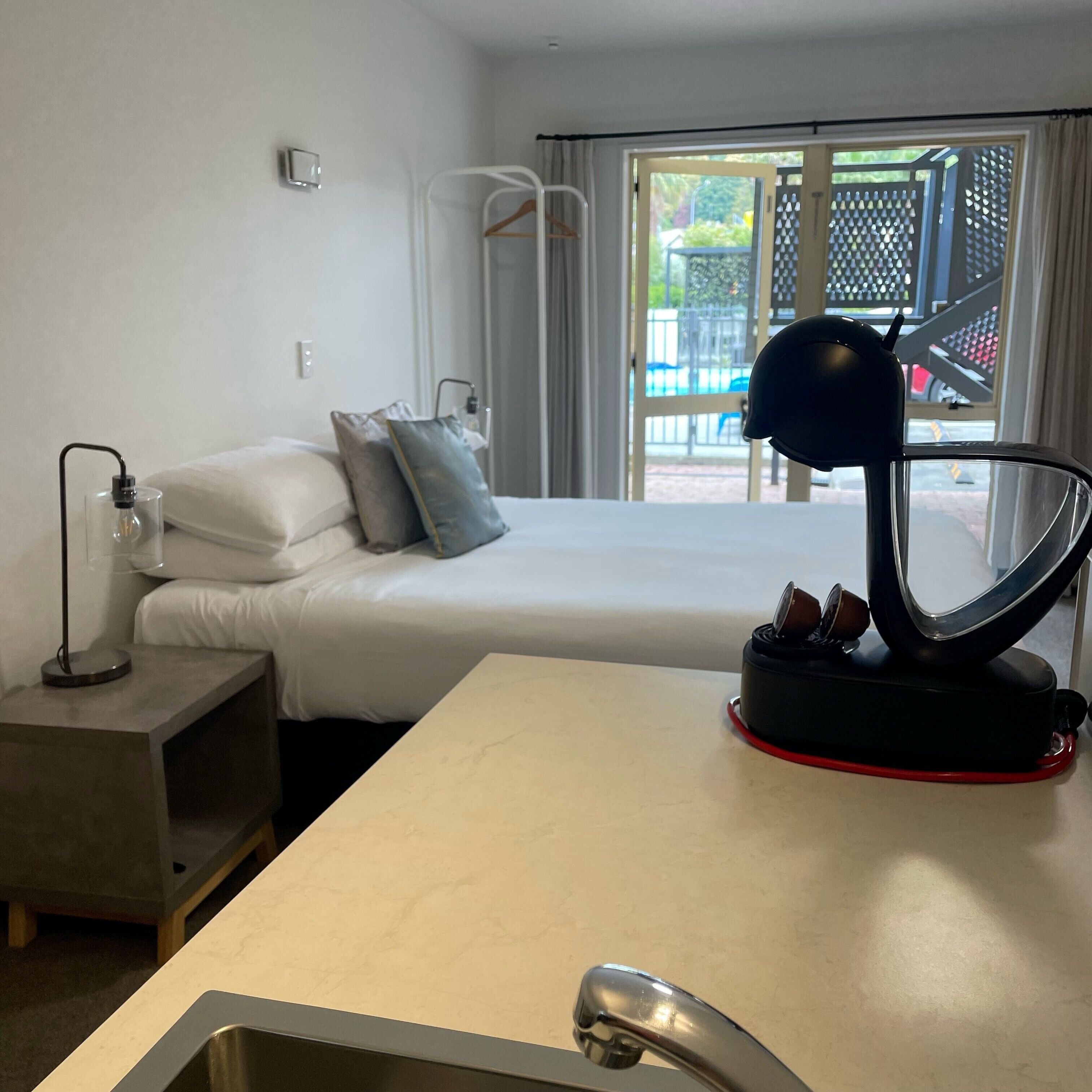 A hotel room with a bed, a bedside table with a lamp, a small table with a coffee pot, and a window with a view of the outside.