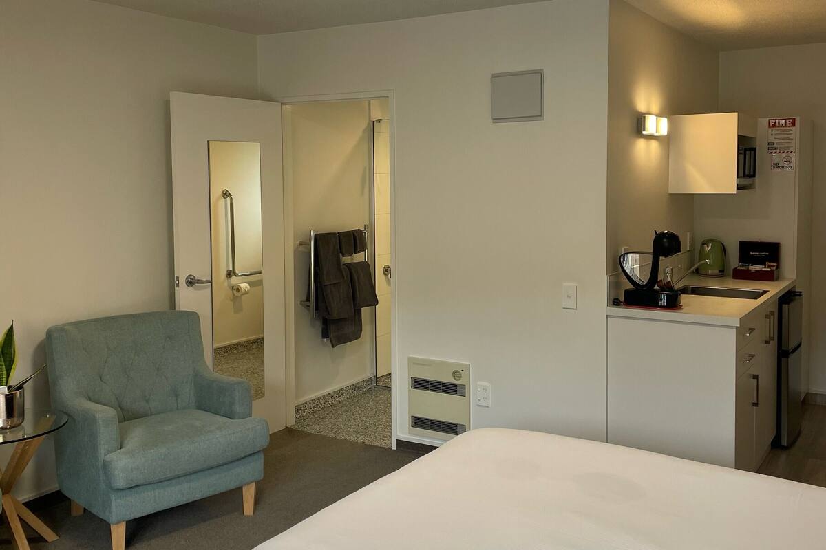 A hotel room with a bed, a chair, a small table with a plant, a kitchenette, and a bathroom visible through an open door.