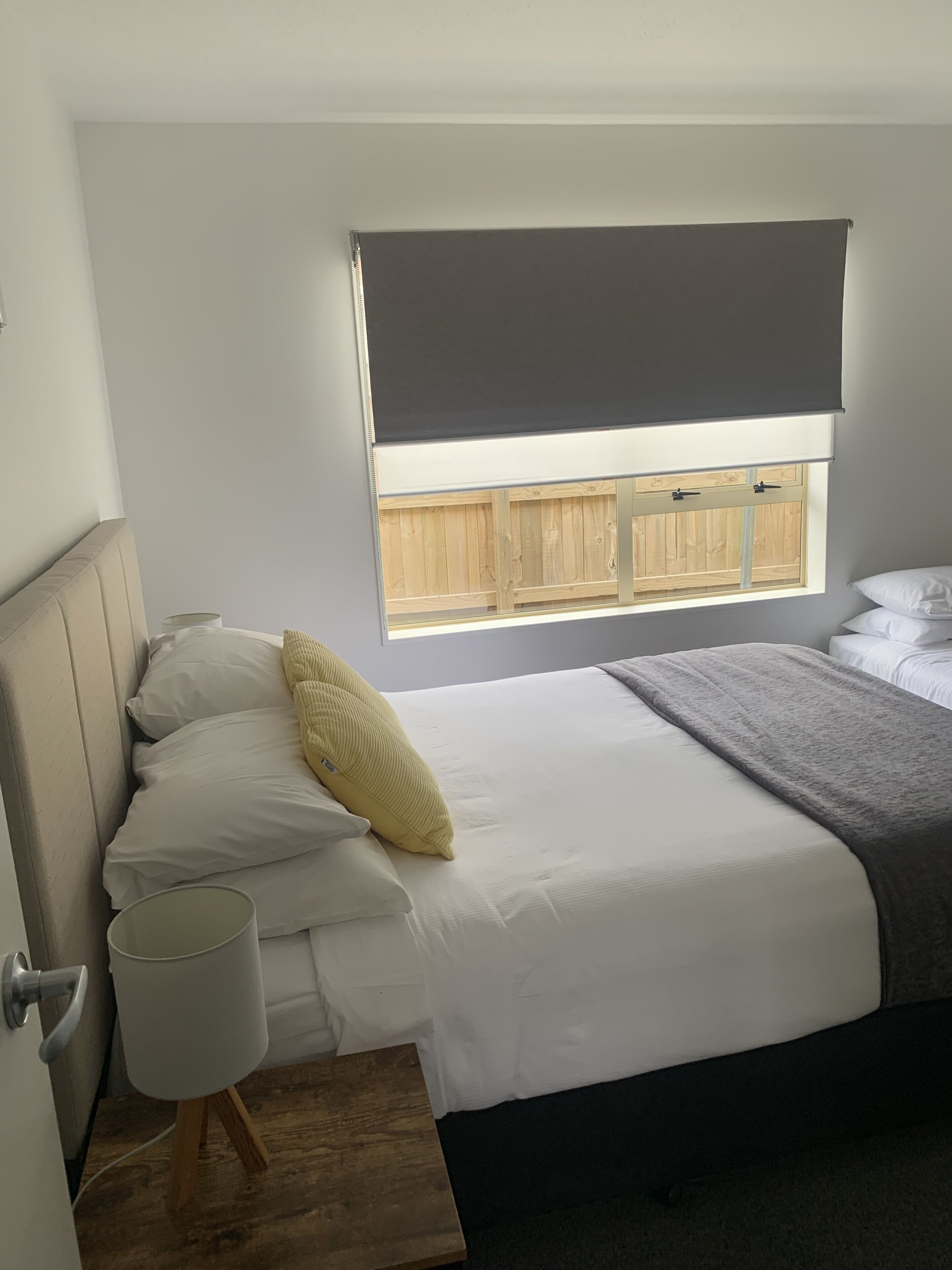 Une chambre à coucher avec un lit, des oreillers, une table de chevet, une lampe et une fenêtre donnant sur une clôture en bois.