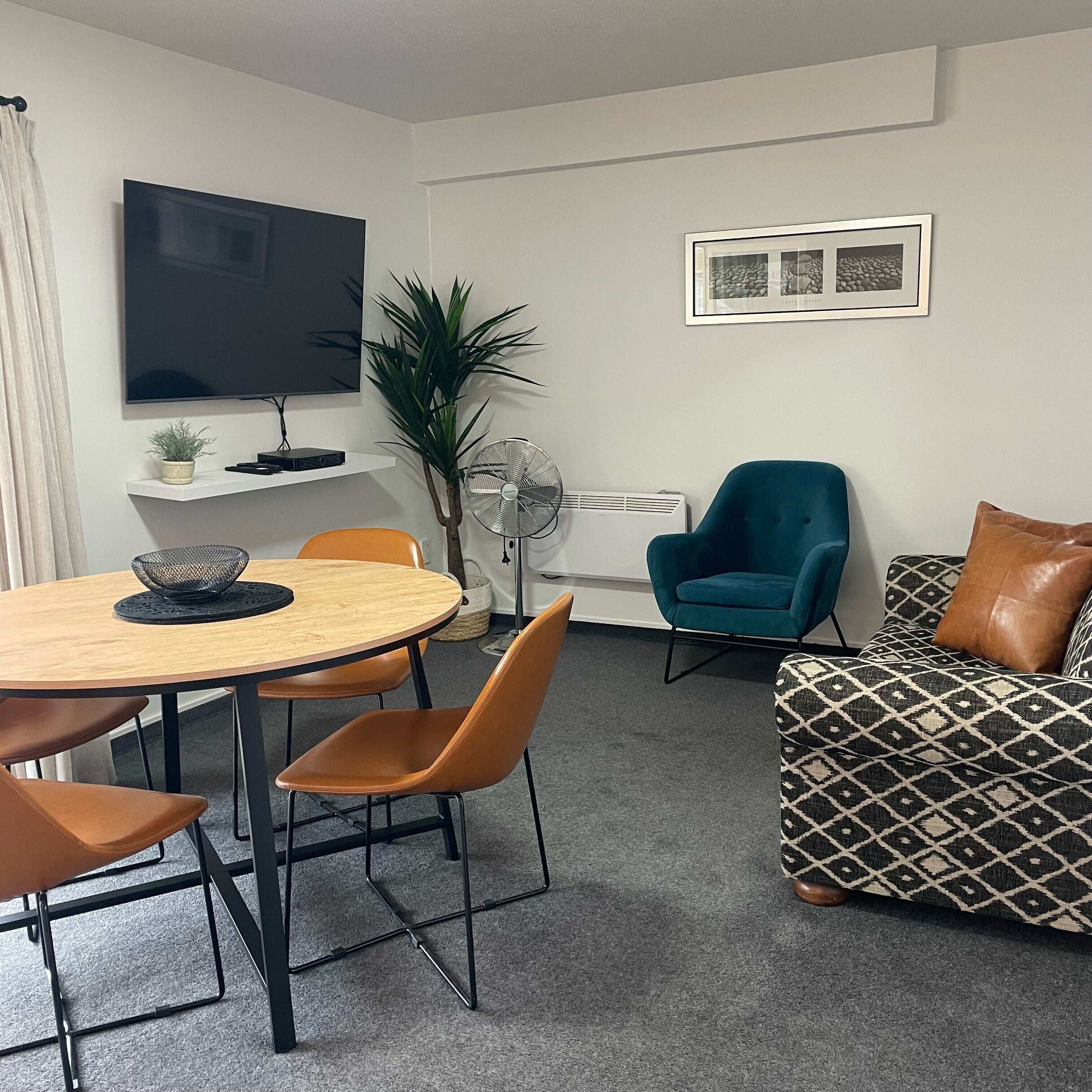 Una sala de estar moderna con una mesa redonda de madera, sillas anaranjadas, un sillón turquesa y un sofá estampado.