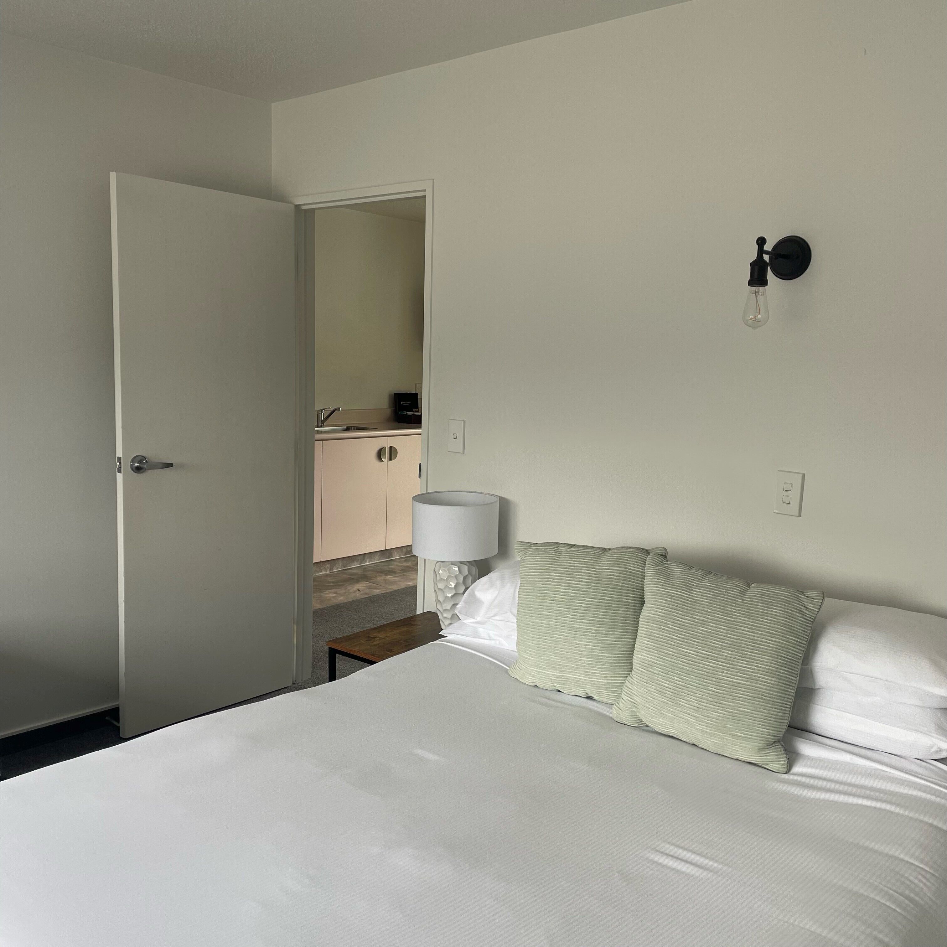 A bedroom with a bed, a nightstand, a lamp, and a view of a bathroom.