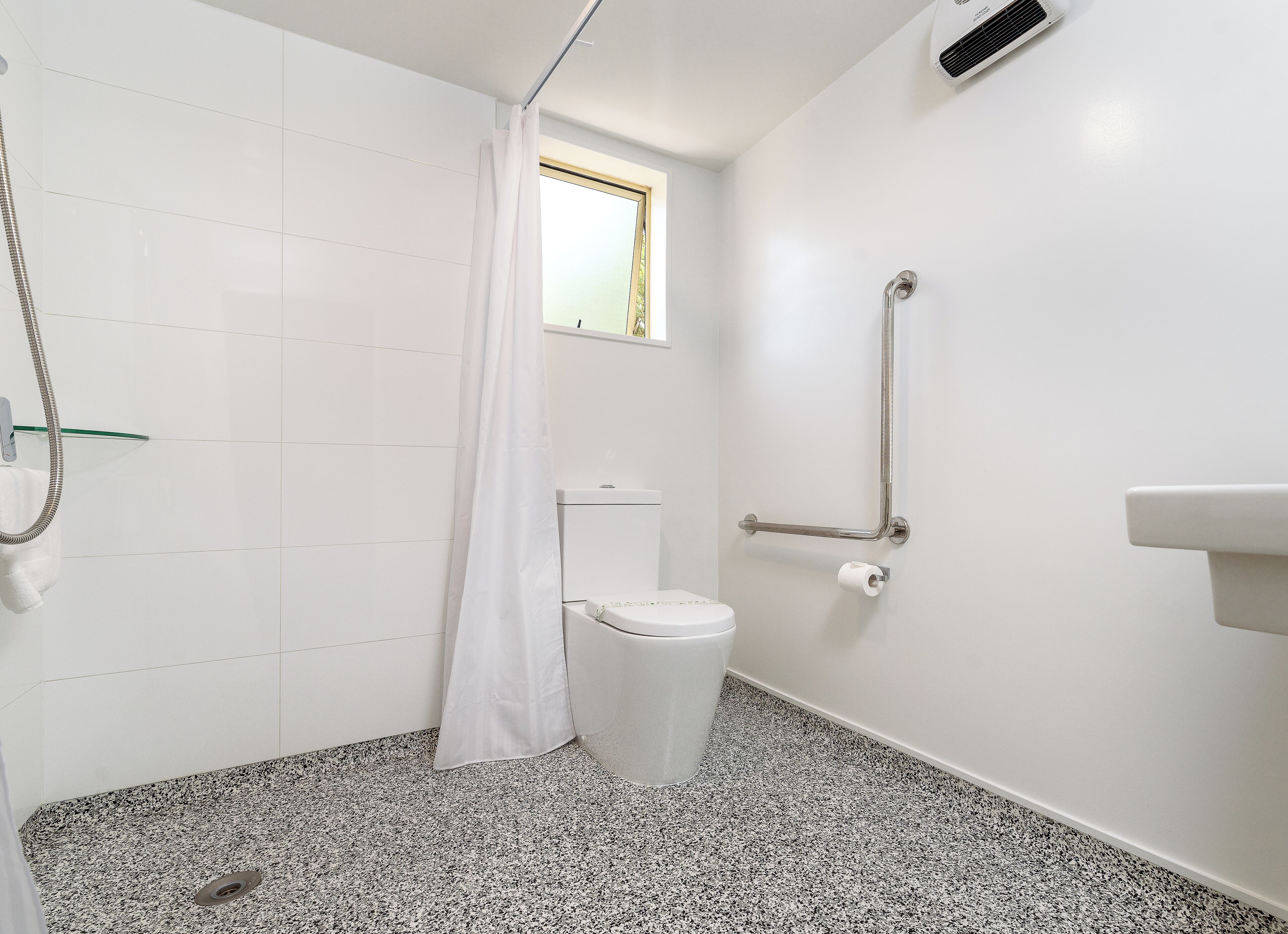 A small, modern bathroom with white tiles, a shower with a glass door, a toilet, and a handrail.