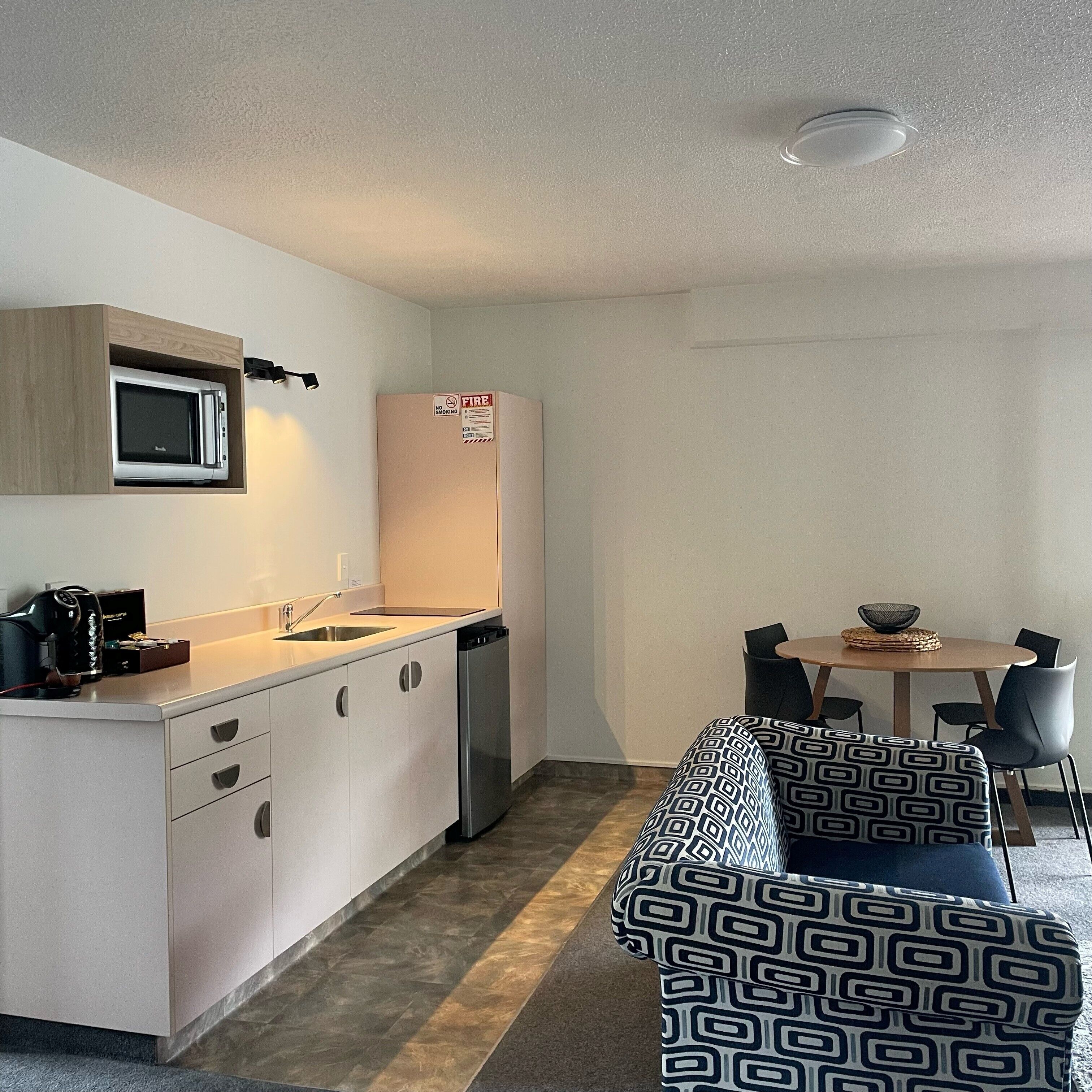 Una cocina compacta con microondas, lavabo y refrigerador, una pequeña mesa de comedor con sillas, y un sofá estampado.