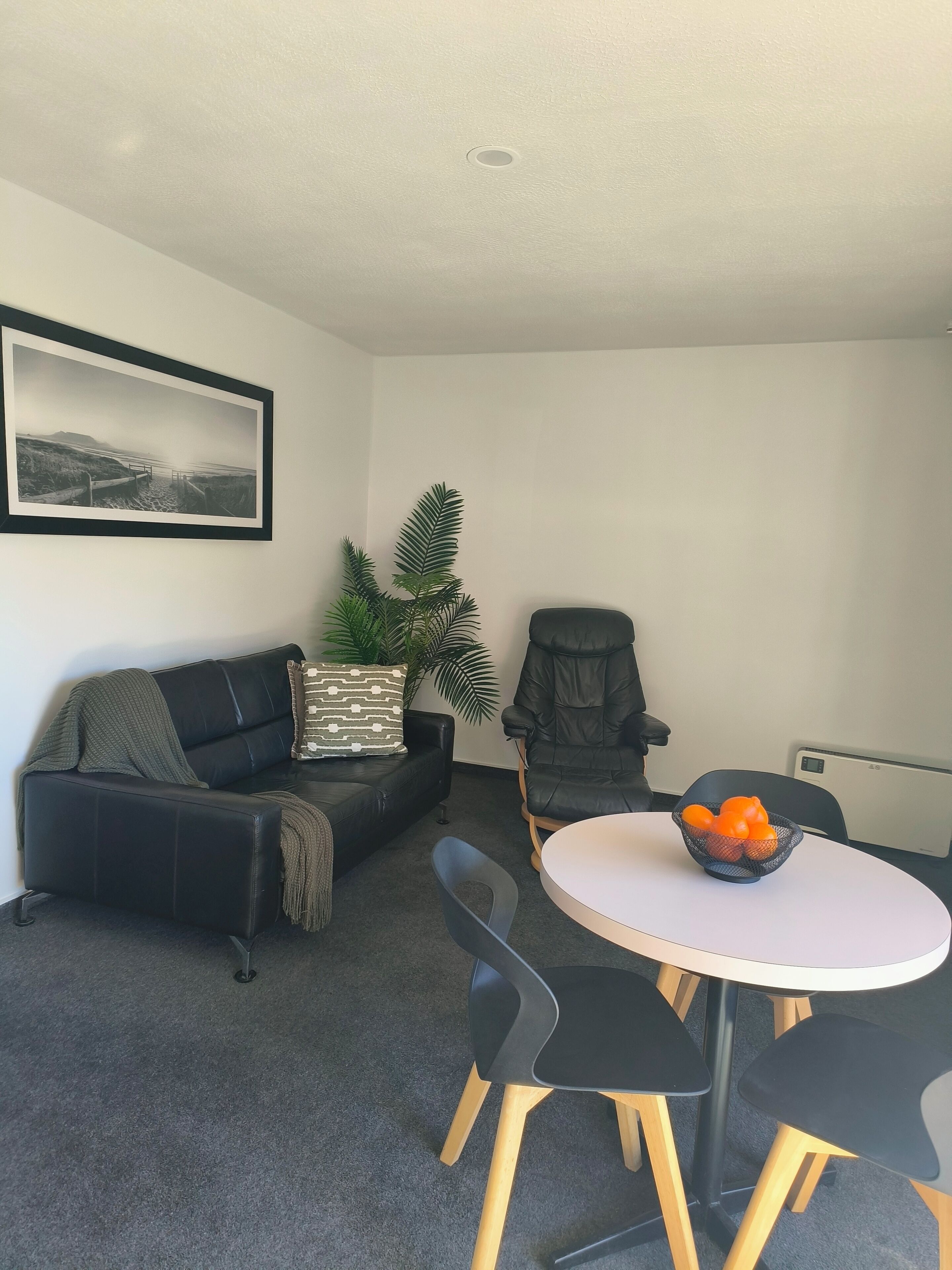 A modern living room with a black leather sofa, a round table with chairs, a black armchair, a potted plant, and a framed photograph on the wall.
