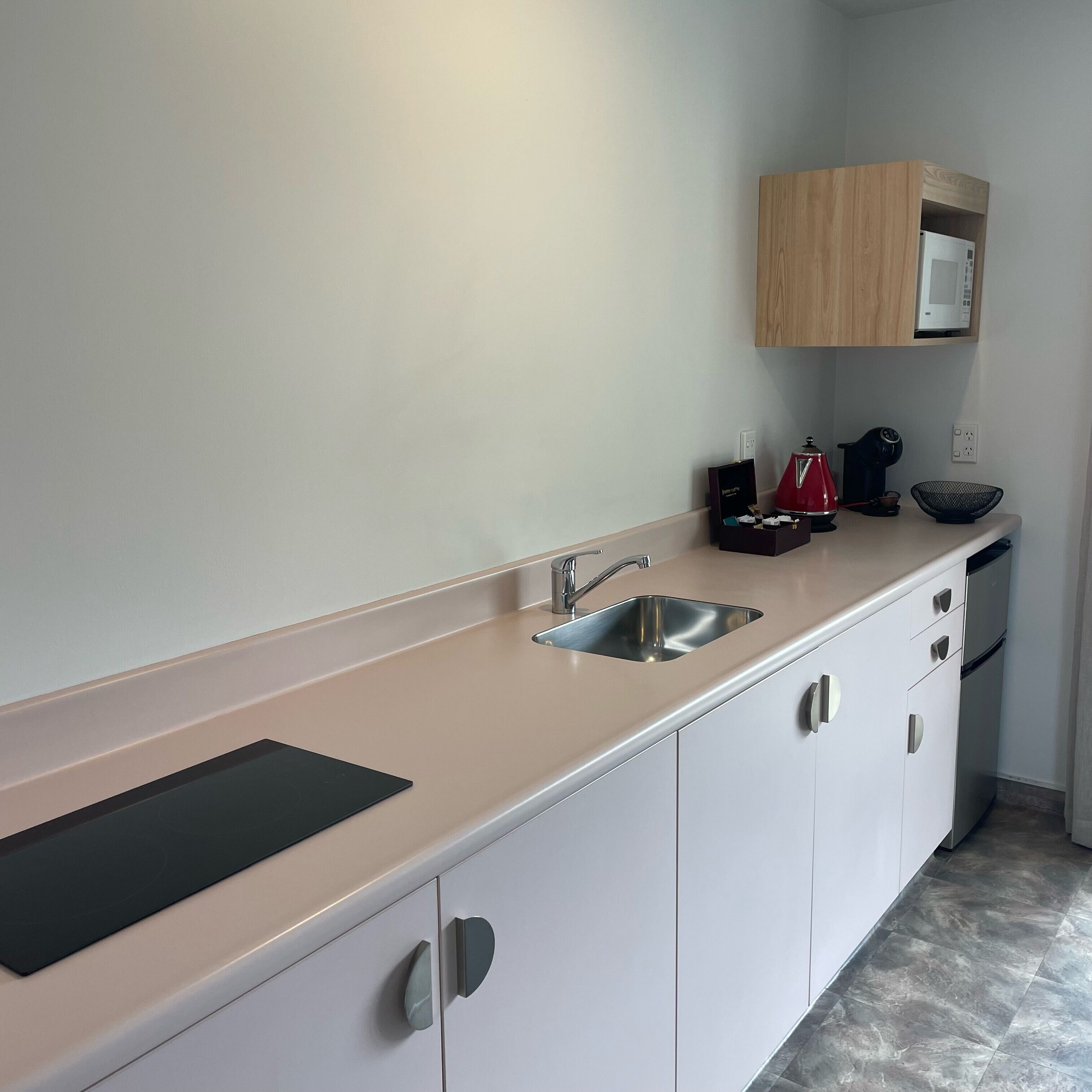 Eine moderne Küche mit Spüle aus Edelstahl, weißen Schränken, einem Holzregal über der Mikrowelle und einem schwarzen Wasserkocher auf der Arbeitsplatte.