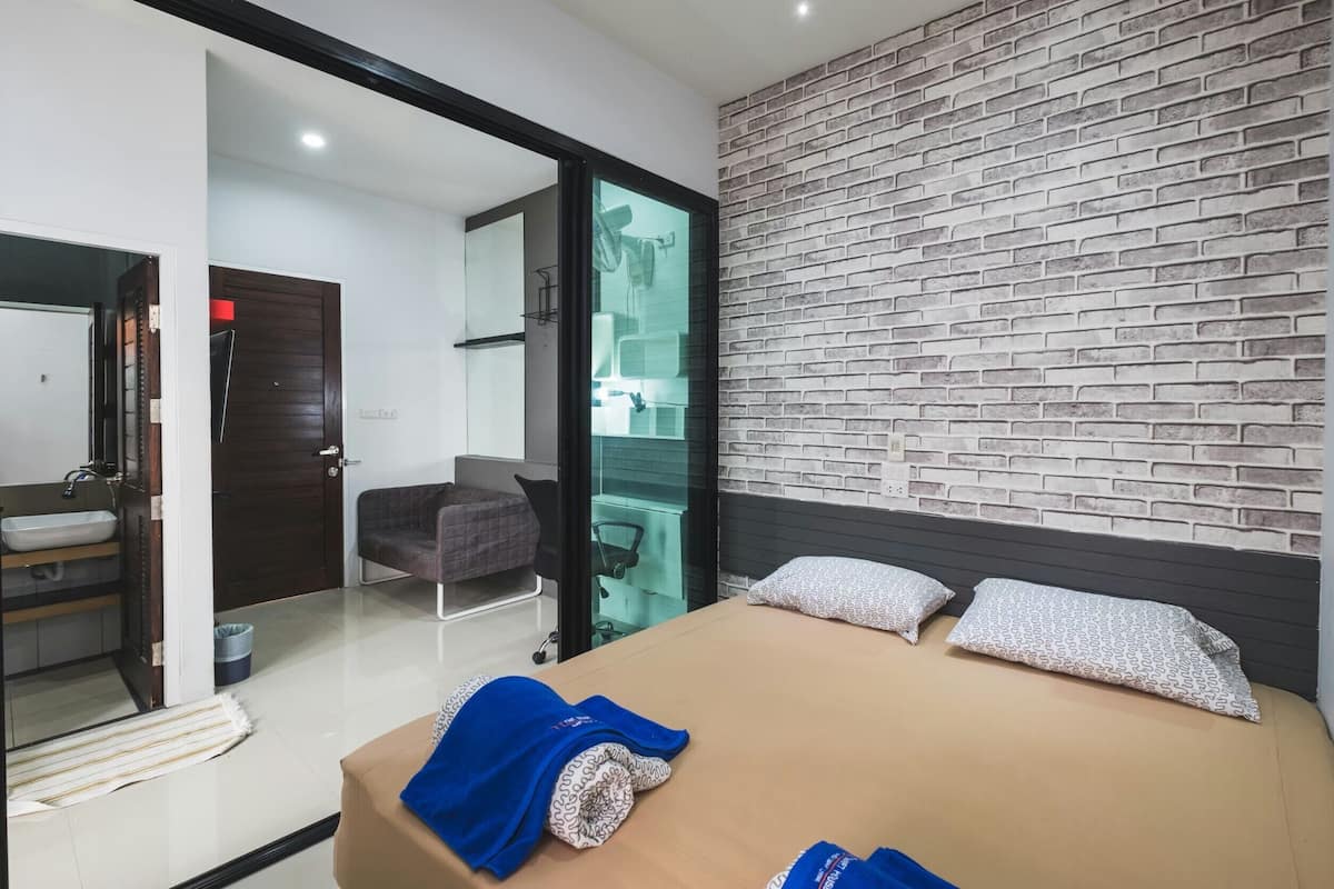 A modern hotel room with a bed, a desk, a chair, a bathroom, and a brick-patterned wall.