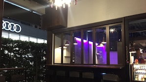 Un área de estar interior con mesas y sillas, un ventanal con iluminación púrpura, y un letrero de Audi visible al fondo.