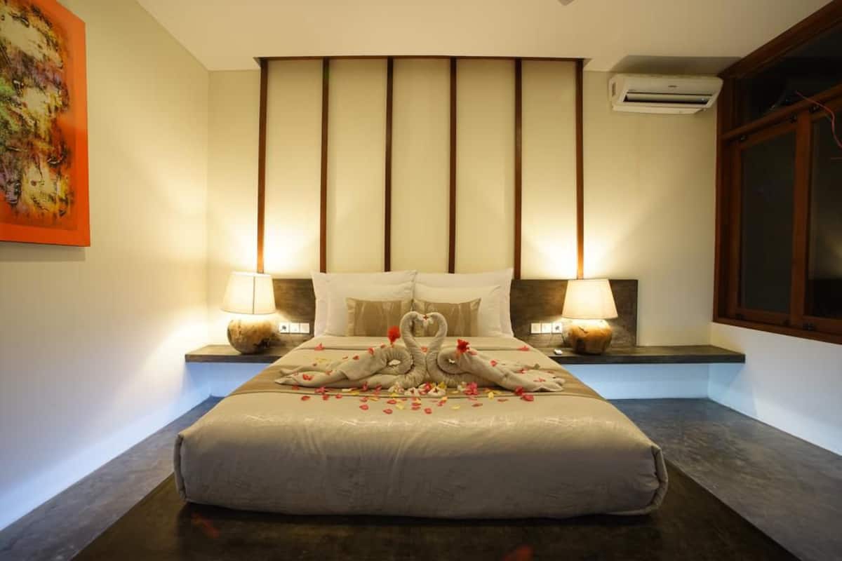 A hotel room with a bed, two bedside lamps, a wall-mounted air conditioner, and a large window.