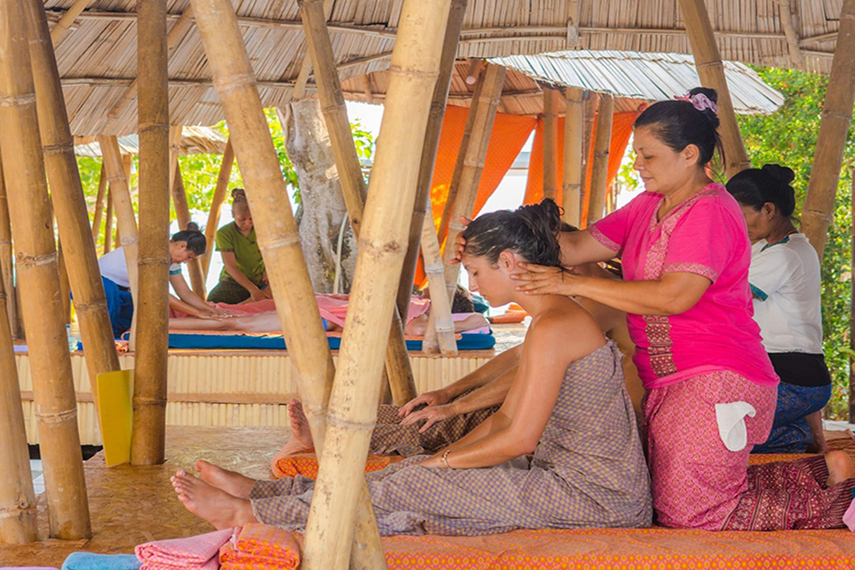 A group of people sitting on mats under a bamboo structure, possibly in a communal area.