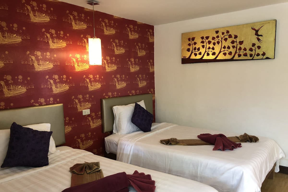 Deux lits simples avec du linge de lit blanc, un mur décoré d’un motif rouge et une œuvre d’art dorée représentant un arbre avec un oiseau.