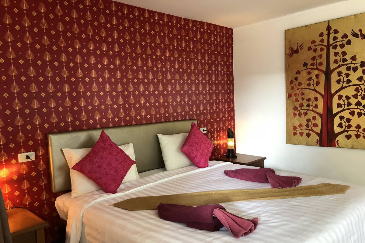 Une chambre d’hôtel avec un grand lit, un papier peint rouge à motifs et un tableau représentant un arbre avec des oiseaux.