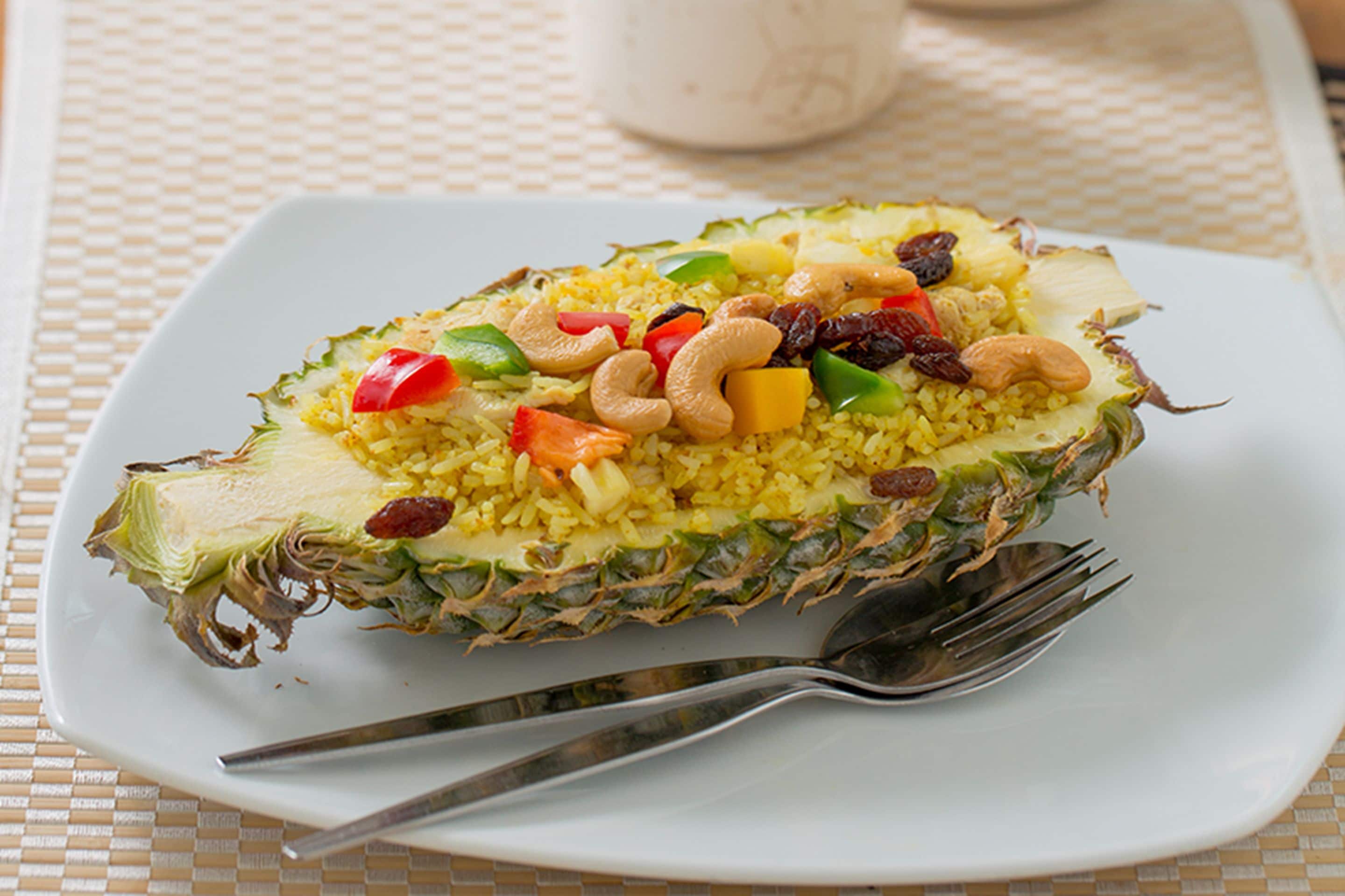 A pineapple filled with yellow rice, red bell peppers, cashews, and raisins on a white plate.