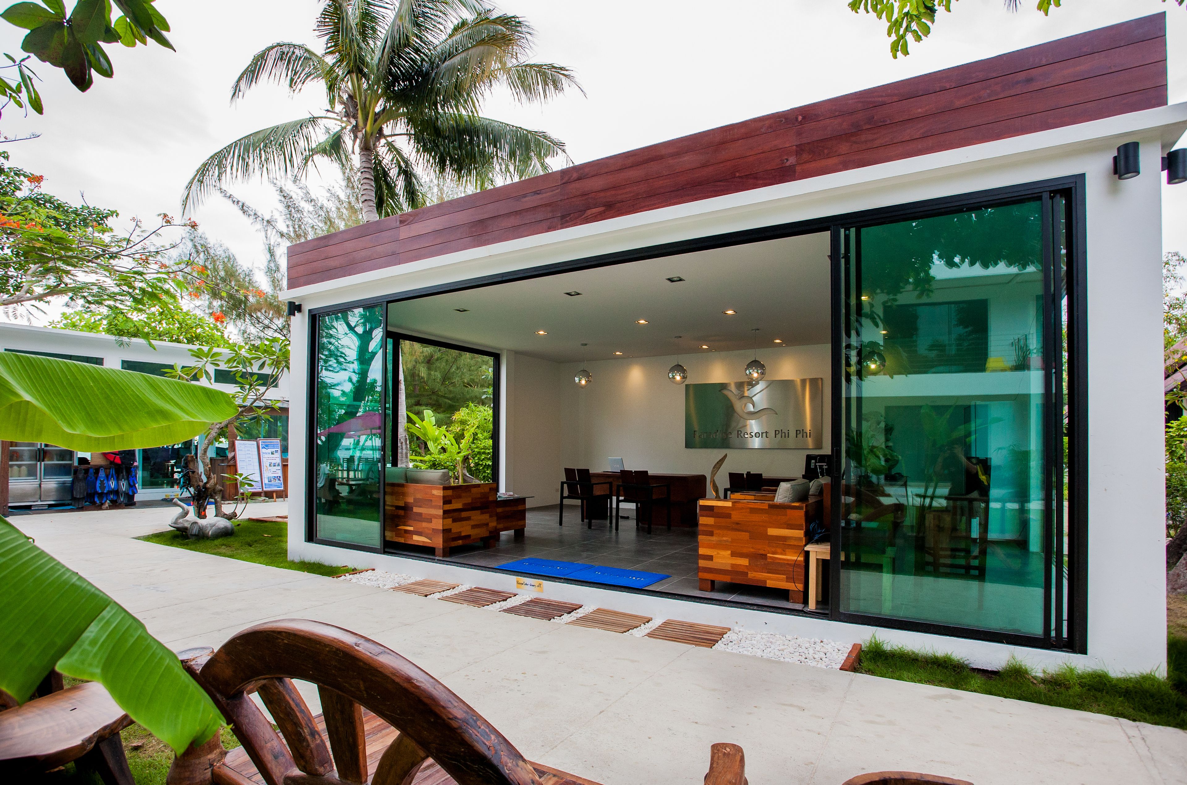Un edificio moderno con amplios ventanales de vidrio, mobiliario de madera y un entorno de jardín tropical.