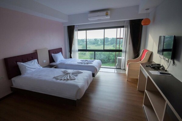 Habitación de hotel con dos camas, un escritorio con silla, televisor y ventana grande con vistas al verdor.