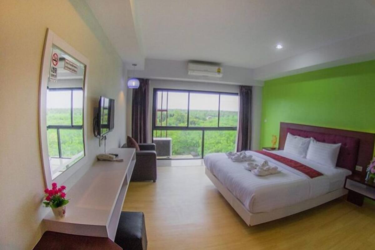 A hotel room with a bed, a desk, a TV, and a window with a view of greenery.